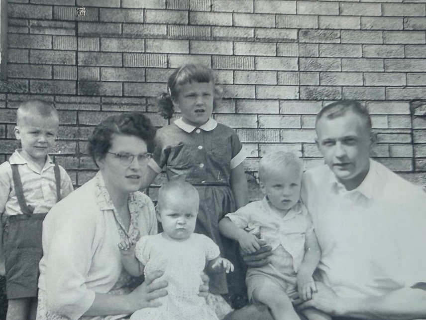 A vintage black and white photo of a family group seated in front of a brick wall. The image includes two adults and four young children, with one child in the mother's lap.